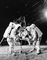 Trainingsfoto aus der Mondhalle in Houston
              (Gebäude 9) mit Armstrong und Aldrin beim
              "Training" für Apollo 11 auf dem Mondboden.
              Rechts oben strahlt ein grosser Scheinwerfer als
              "Sonne" mit fotografischem Strahleneffekt.